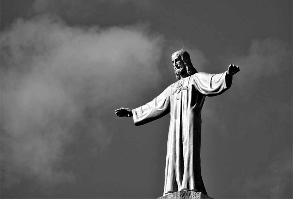 Statue du Christ les bras grands ouverts en signe d'accueil, représentée en noir et blanc sur un fond de ciel nuageux, symbolisant l'ouverture du chemin de la foi à tous.